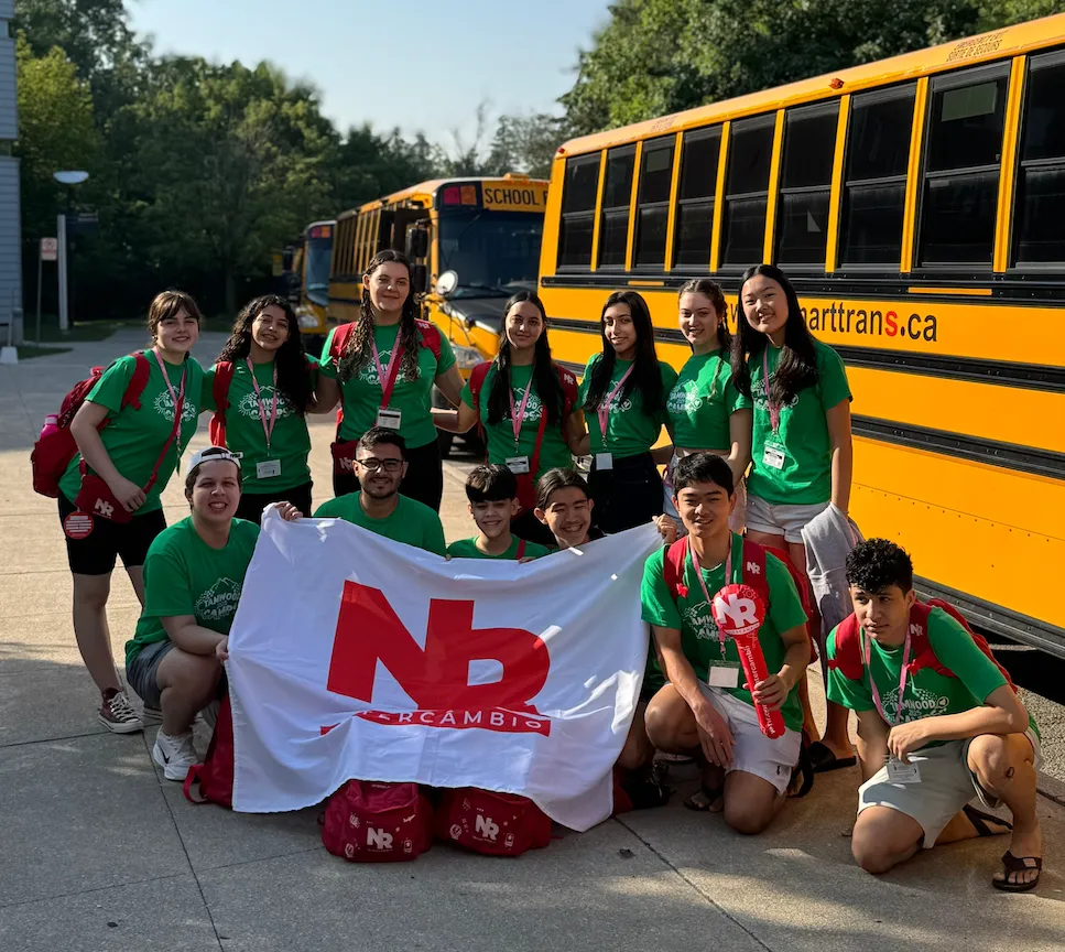 Grupo de alunos em intercâmbio posando com uma bandeira ao lado de ônibus escolares amarelos